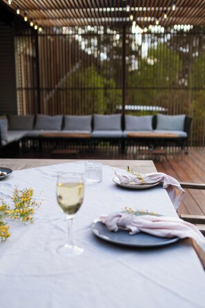 A dining table set with a glass of white wine, a plate with food, and a napkin. There is a bench with cushions in the background.