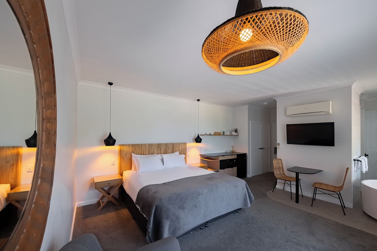 A modern hotel room with a large bed, bedside tables, a desk, a chair, a small table, a TV, and a bathroom with a bathtub.
