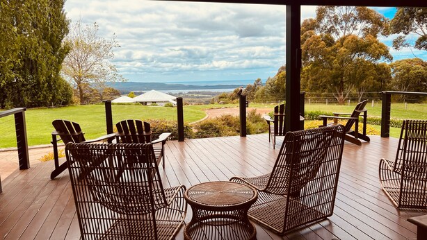 A covered outdoor seating area with wicker chairs and a table, overlooking a grassy landscape with trees and a distant view of hills. - Chimes Spa Retreat (Shadforth)