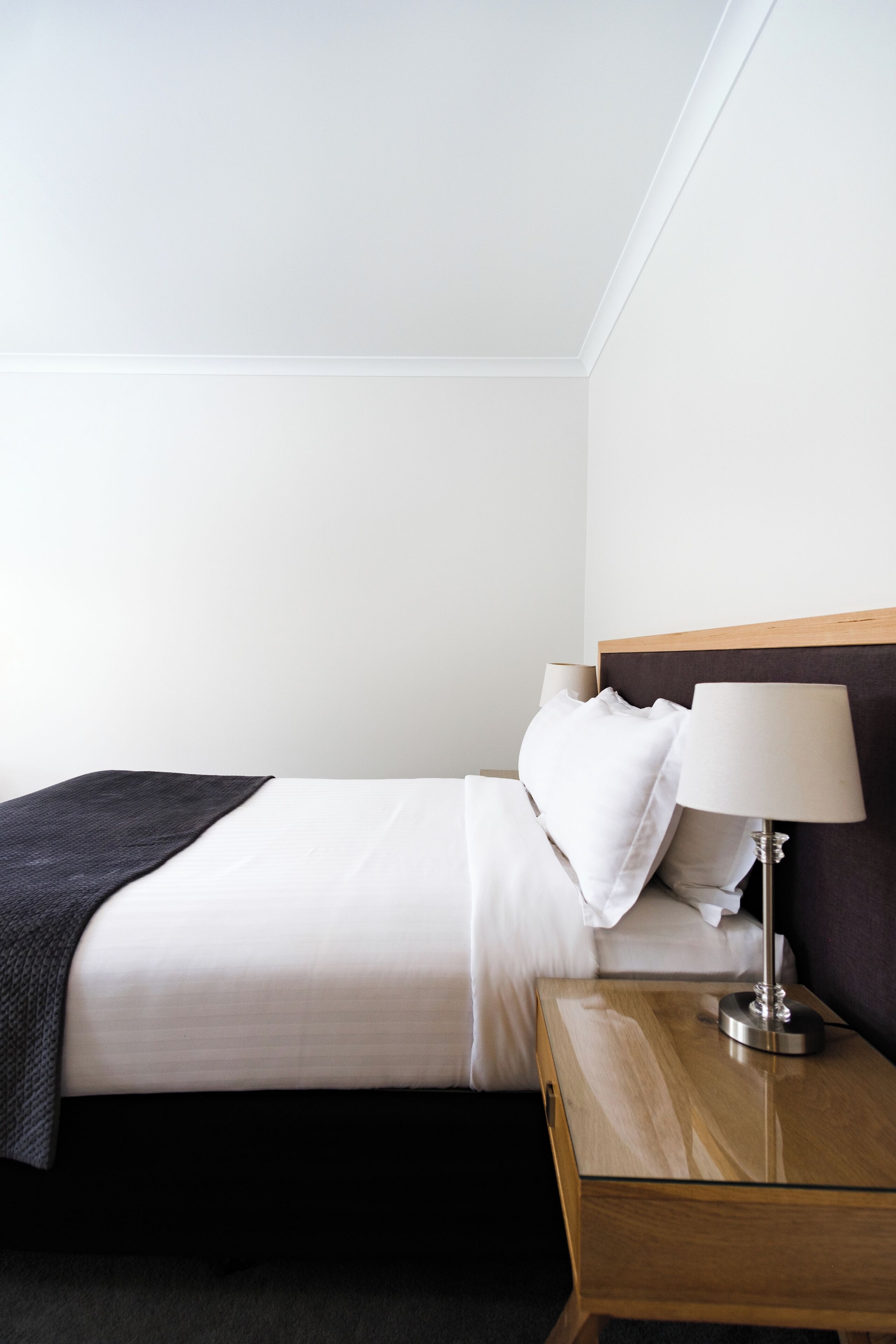 Una cama bien tendida con sábanas blancas, un plaid oscuro y una mesita de noche con una lámpara.