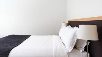 Una cama bien tendida con sábanas blancas, un plaid oscuro y una mesita de noche con una lámpara.