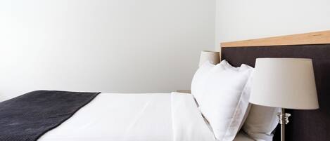 Cama bem arrumada com lençóis brancos, um cobertor escuro e uma mesa de cabeceira com abajur.