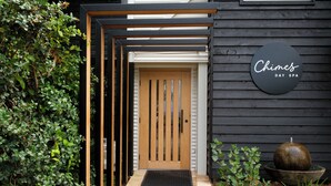A modern entrance with a wooden door, a black wall with a circular sign, and a stone planter with a spherical object. - Chimes Spa Retreat (Shadforth)