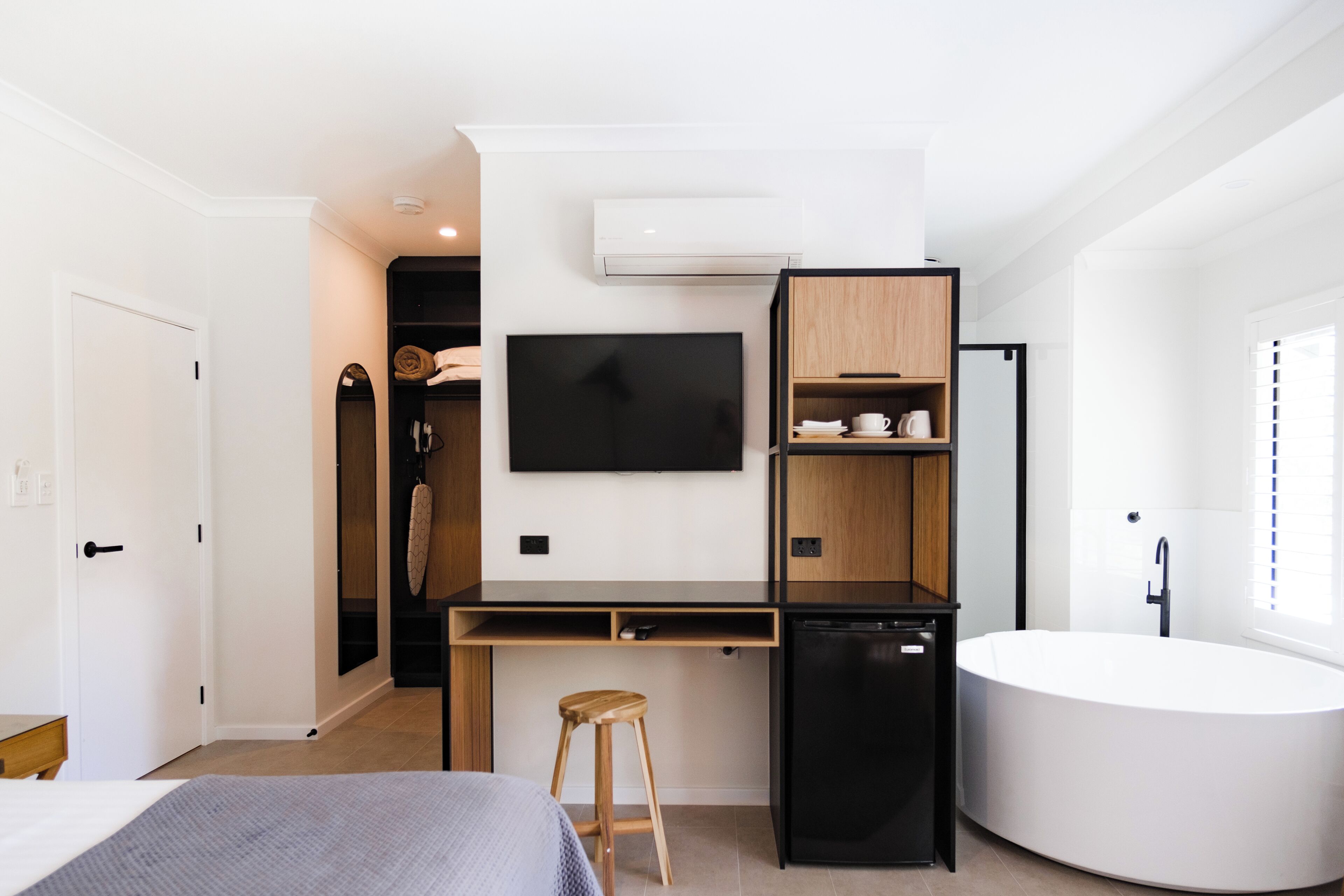 A modern hotel room with a bed, a desk with a chair, a flat-screen TV, a built-in dishwasher, a freestanding bathtub, and a wardrobe.