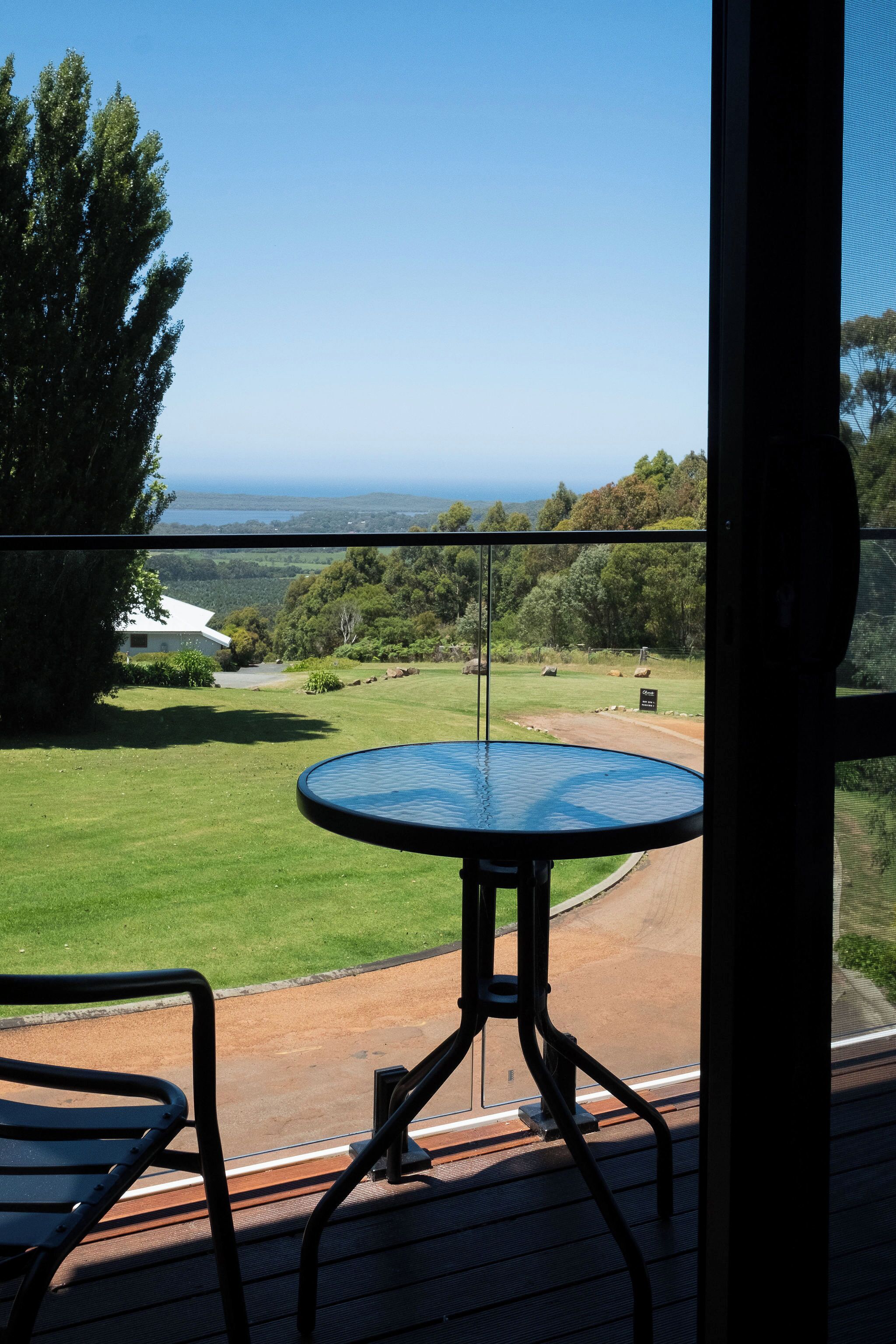Un balcón con mesa y sillas, con vistas a una zona verde con árboles y una vista distante del océano.