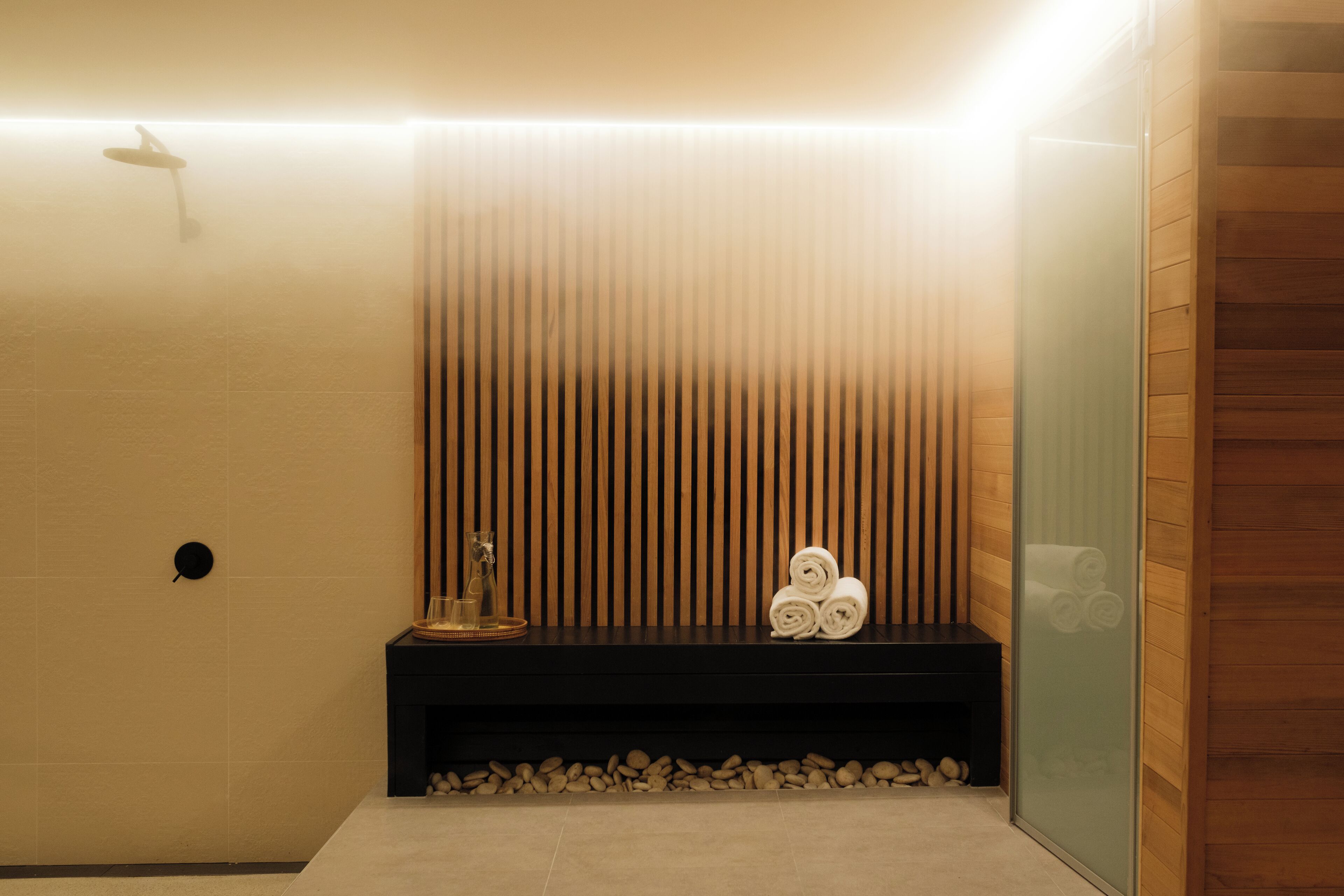 Un baño moderno con un panel de pared de madera, un banco negro con toallas y un box de ducha de cristal.