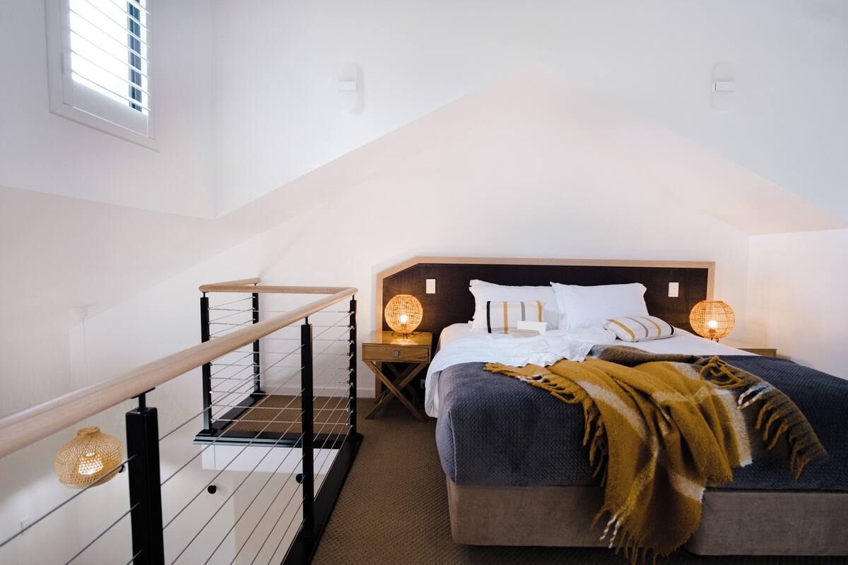 A bedroom with a sloped ceiling, a wooden staircase, a bed with a headboard, two bedside lamps, and a window with blinds.