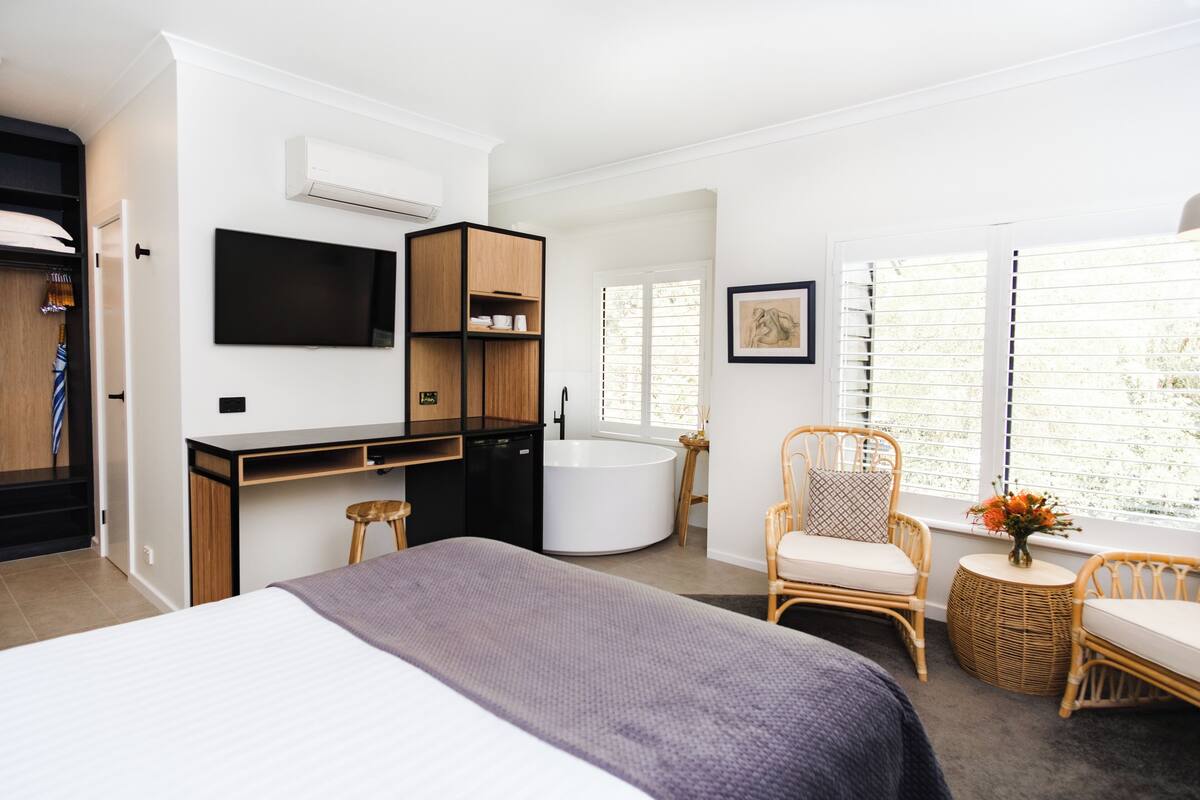 A modern hotel room with a bed, a desk, a chair, a small table, a TV, a bathtub, and large windows.