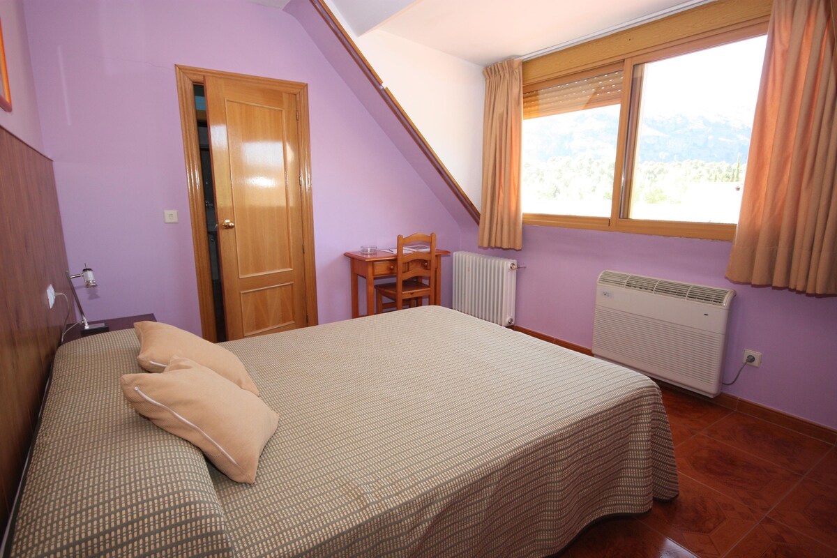 A bedroom with a bed, a chair, a table, a window with curtains, and a radiator.
