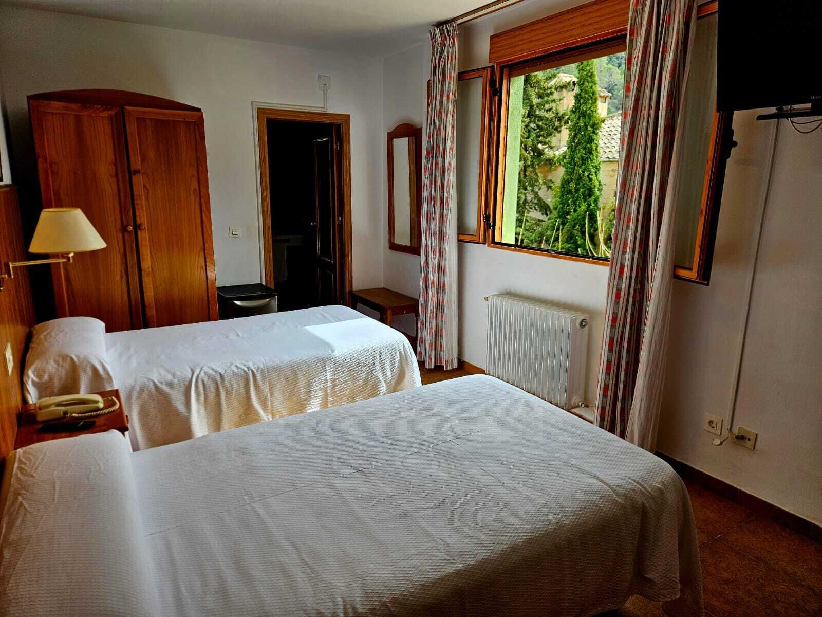 A hotel room with two beds, a wooden wardrobe, a radiator, and a window with curtains.