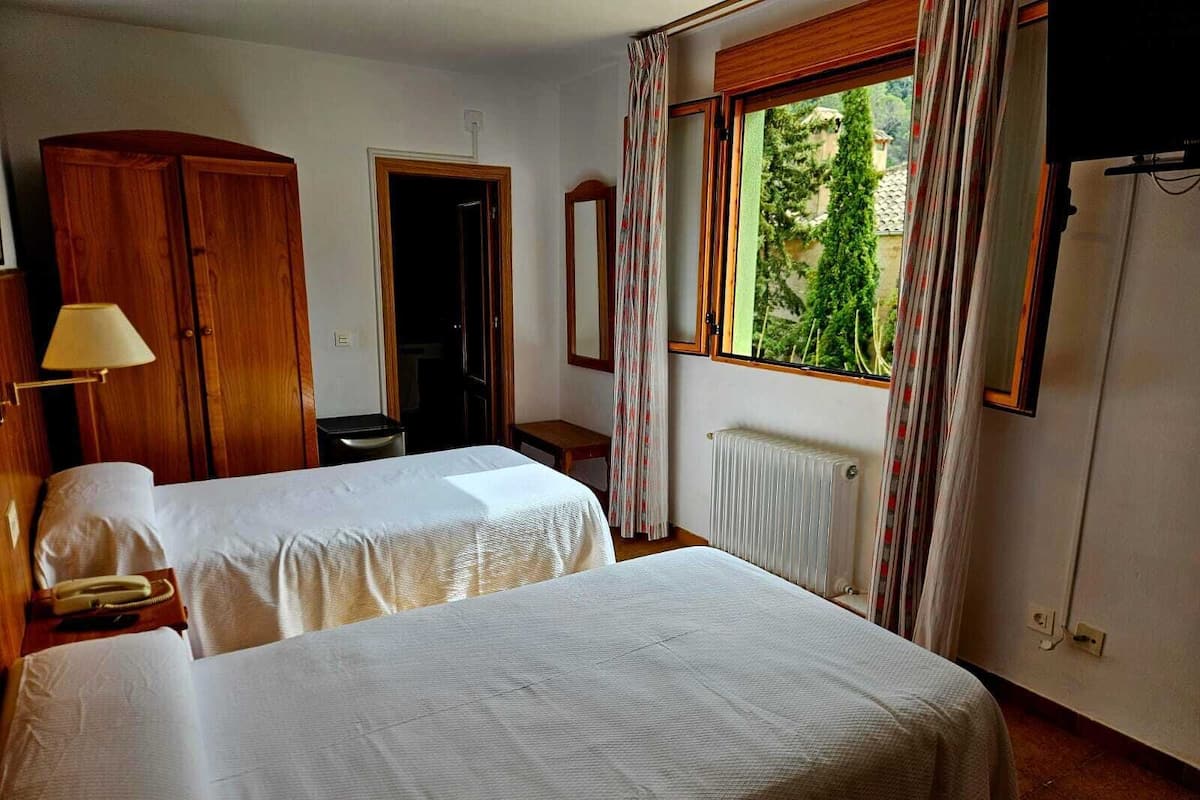 A hotel room with two beds, a wooden wardrobe, a radiator, and a window with curtains.