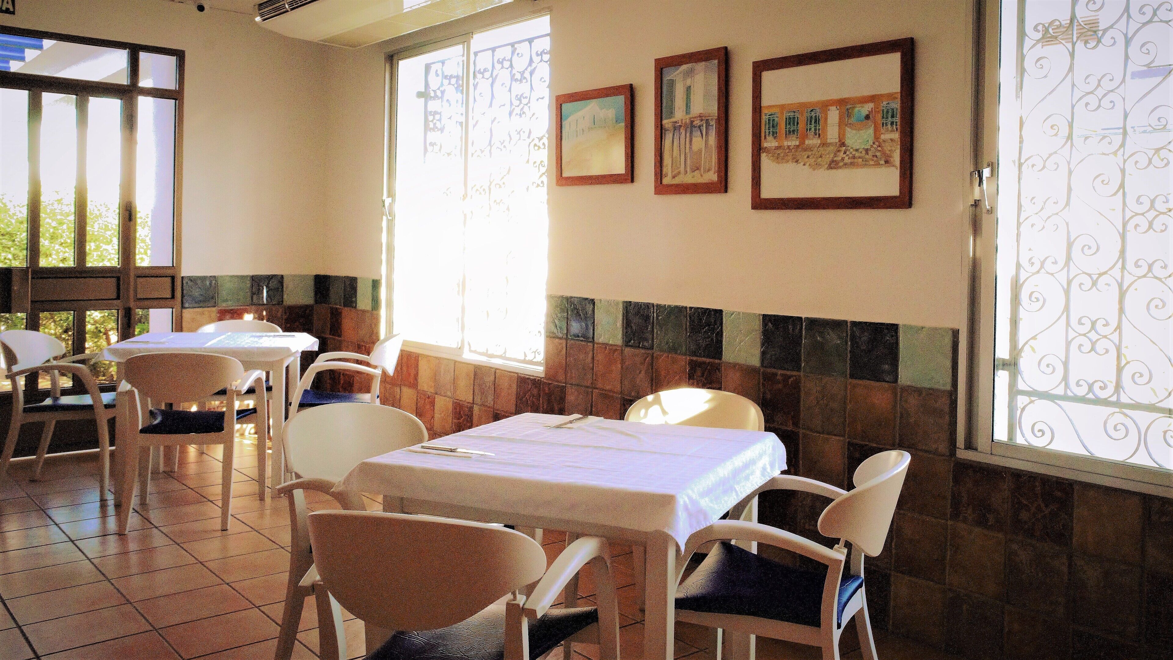Un interior de restaurante con mesas y sillas, ventanas amplias con celosías de hierro decorativas y paredes de azulejos.