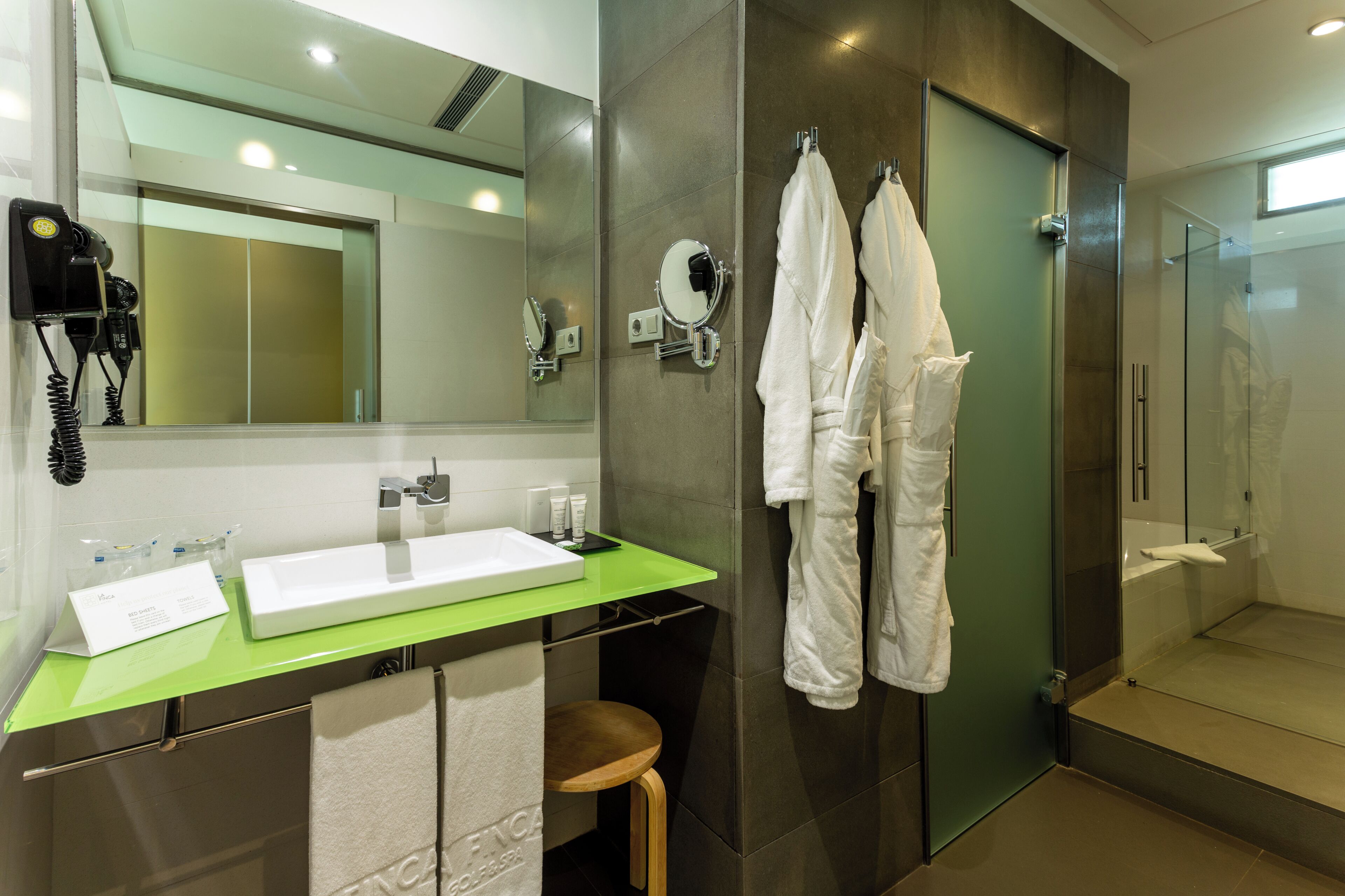 Een moderne badkamer met een groen aanrecht, een witte wastafel, een spiegel, een haardroger, en twee witte badjassen die aan een rek hangen.