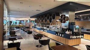 A modern cafe with a long counter, black chairs, and a variety of food and drink options. - La Finca Resort (Algorfa)