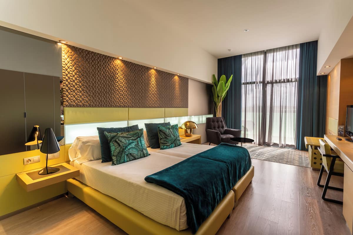 A modern hotel room with a bed, bedside table, lamp, chair, desk, and large window.