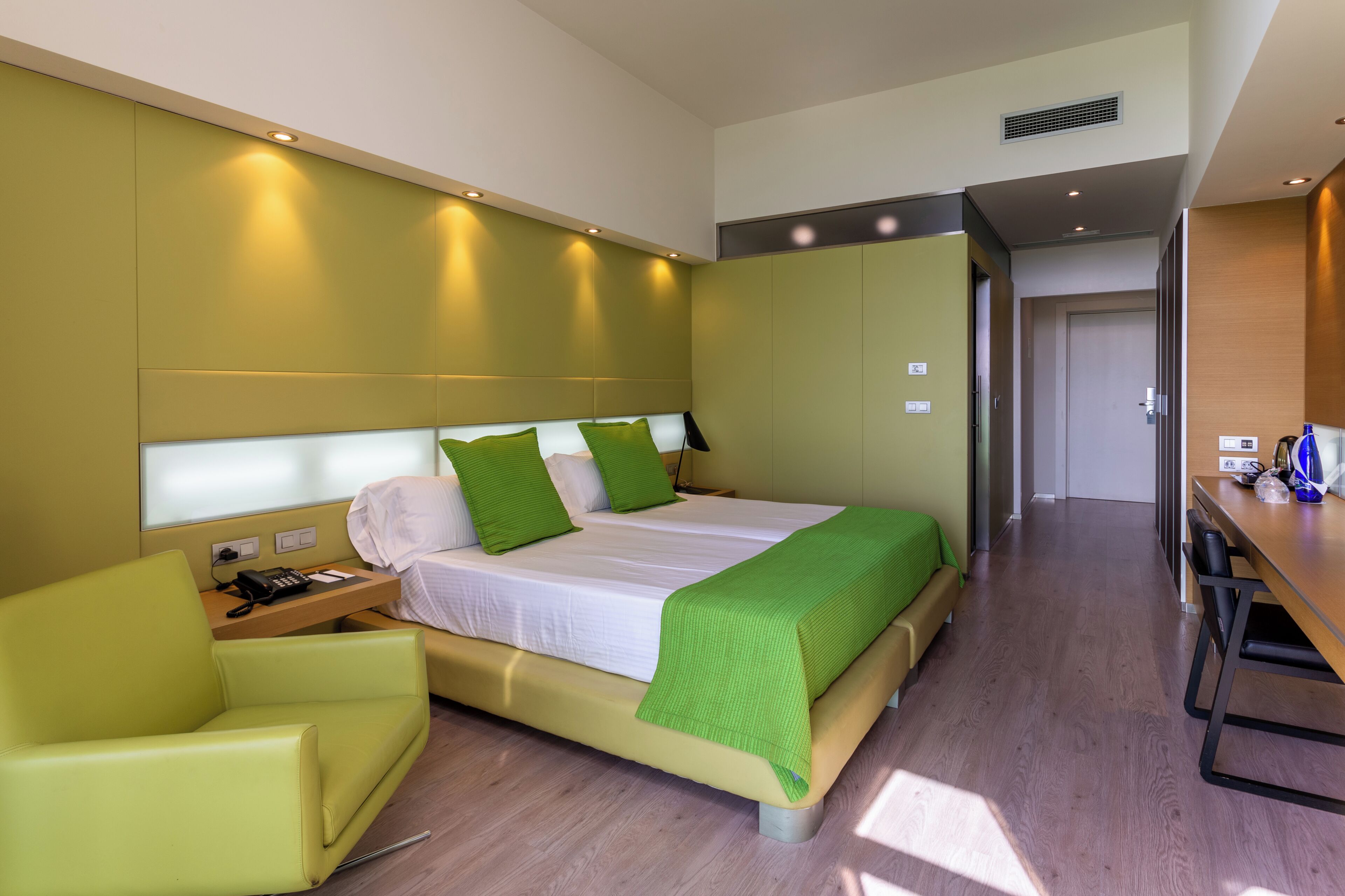 Een moderne hotelkamer met een bed, een groene fauteuil, een bureau en een houten vloer.