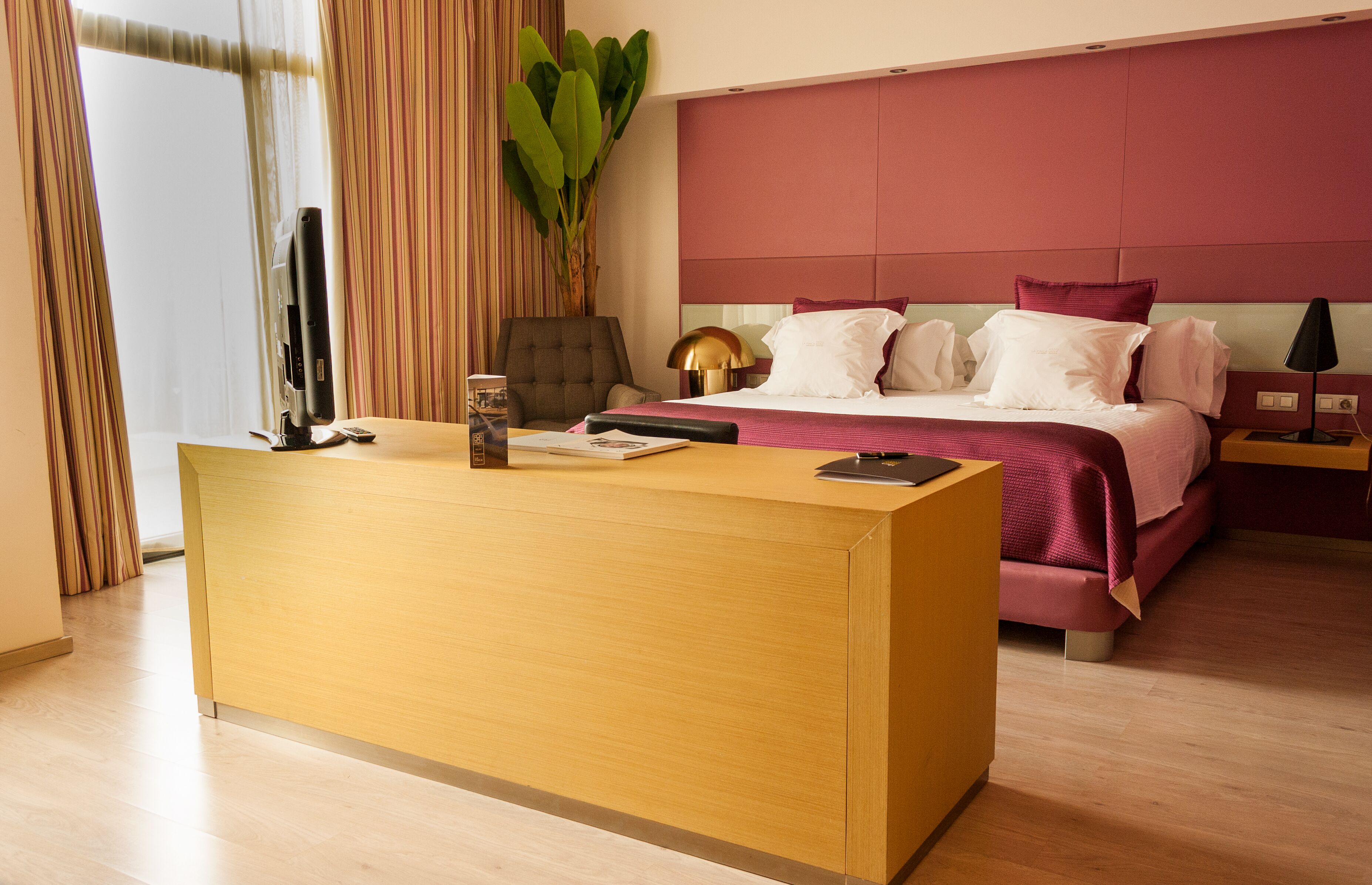 Habitación de hotel con un mostrador de recepción de madera, una cama con cabecera roja, una planta grande en maceta y una mesita de noche con lámpara.