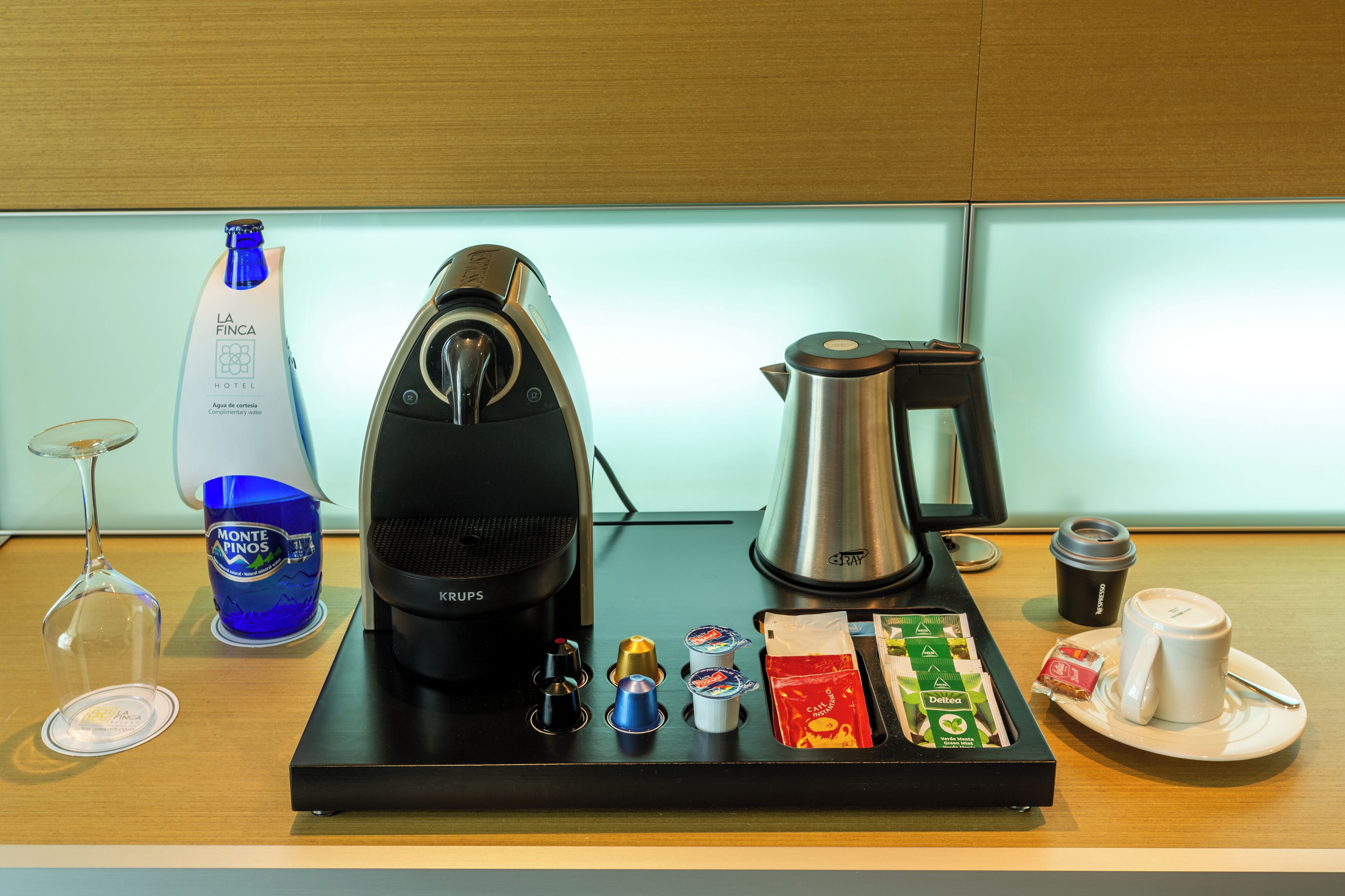 Una estación de café con cafetera, hervidor, vaso de agua, botella de agua, copa de vino y taza de café.