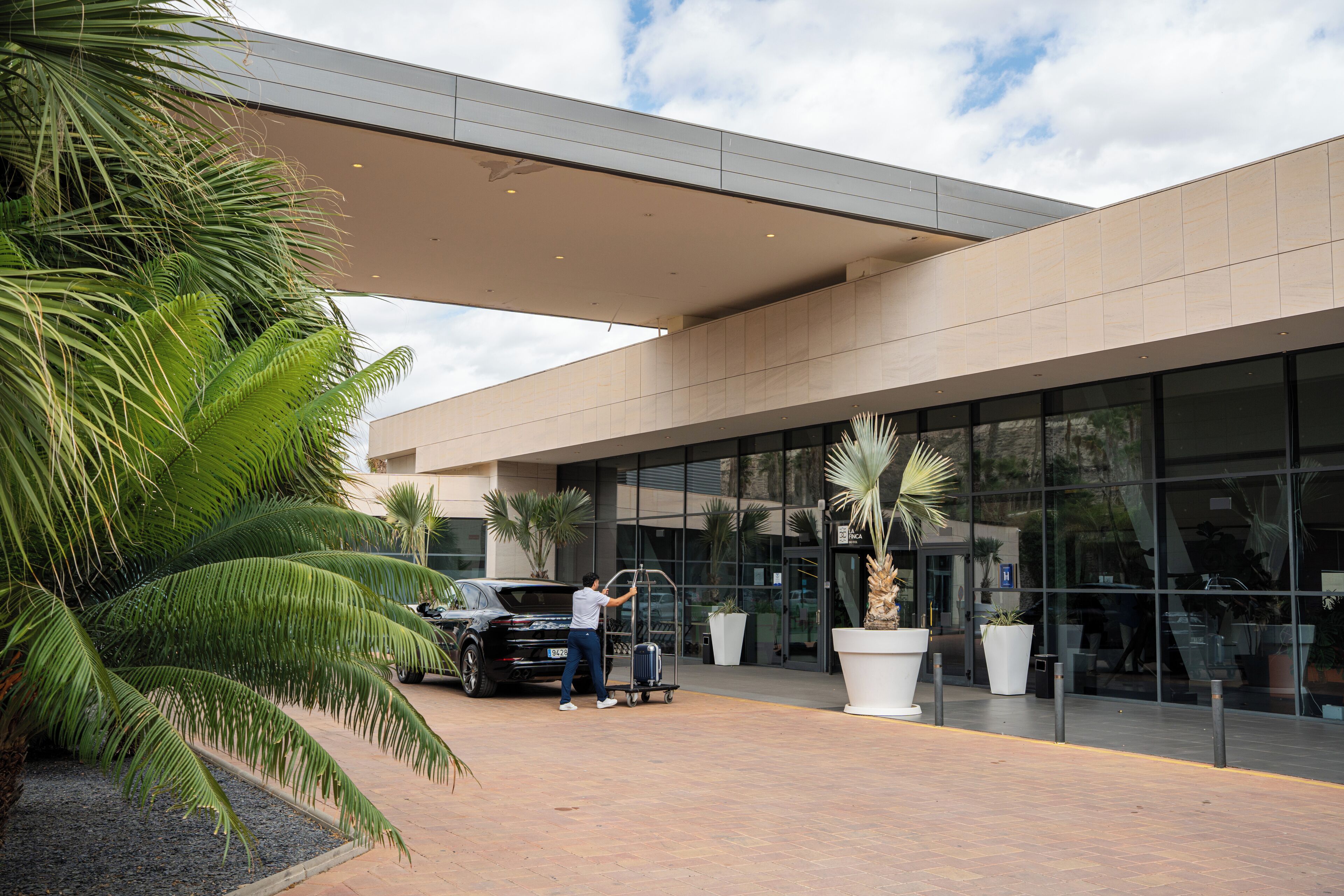 Entrada moderna de un hotel con un paseo techado, palmeras y un coche negro estacionado afuera.