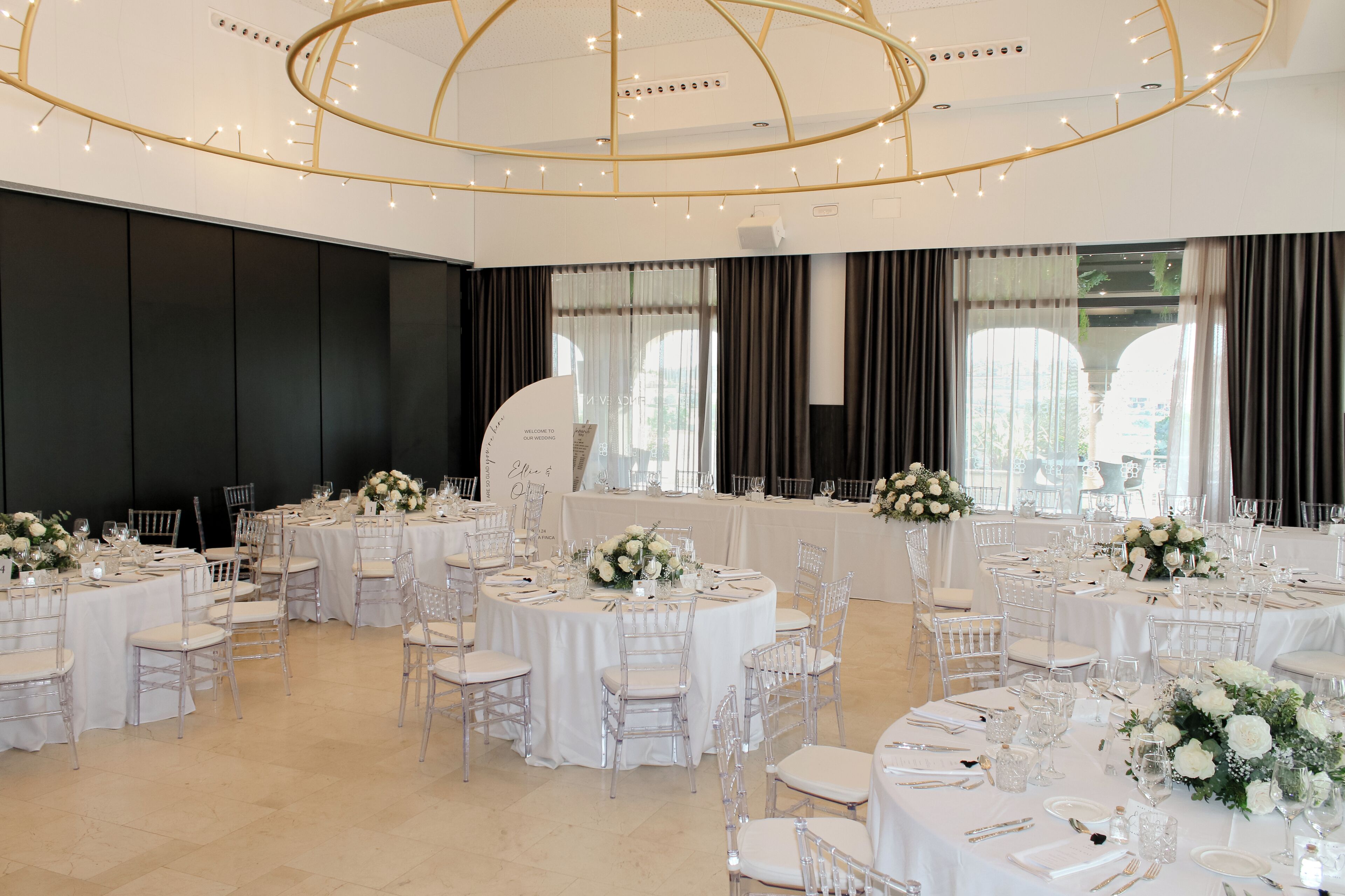 A banquet hall with round tables, white tablecloths, and clear chairs.