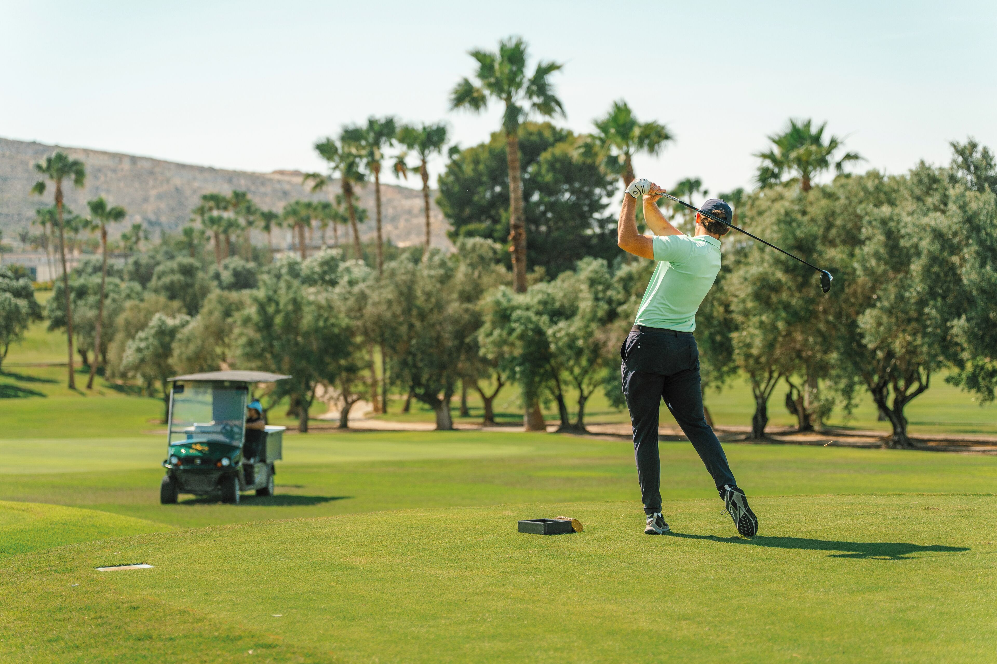 Un golfista en pleno balanceo en un campo de golf, con un carrito y árboles al fondo.