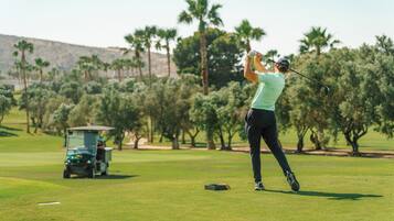 Un golfista en pleno balanceo en un campo de golf, con un carrito y árboles al fondo.