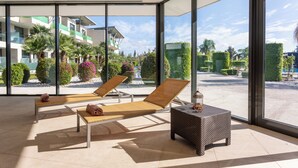 Habitación iluminada por el sol con dos sillones, una mesita y ventanas amplias que dan a un jardín ajardinado y edificios.