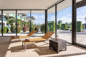 A sunlit room with two lounge chairs, a small table, and large windows offering a view of a landscaped garden and buildings. - La Finca Resort (Algorfa)