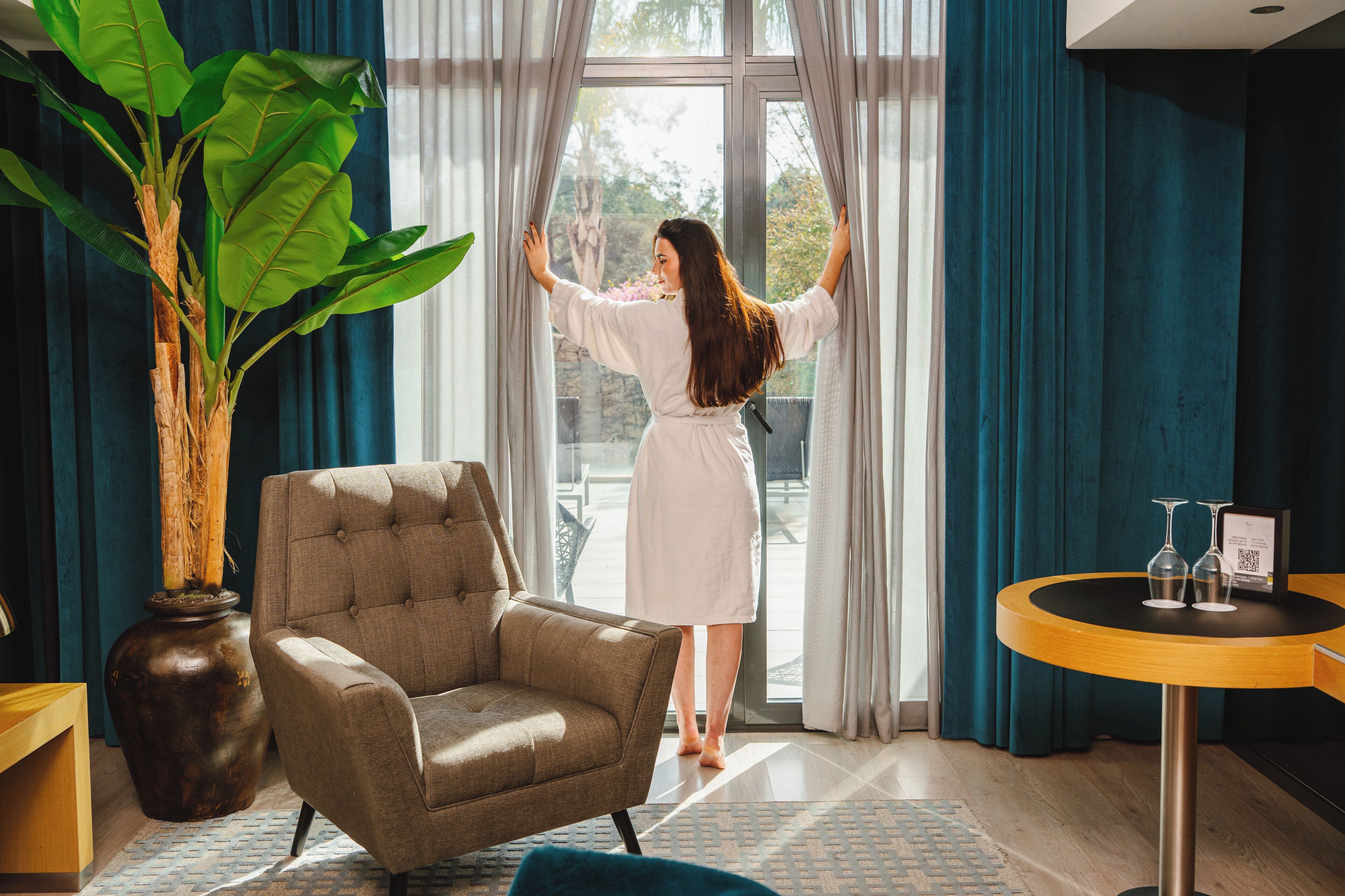 Een kamer met een grote potplant, een fauteuil met een bekleding, een ronde houten tafel met glazen en een persoon die gordijnen rechtzet.