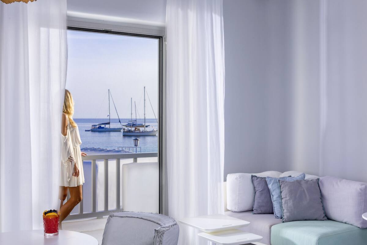 A room with a view of sailboats at the marina, a balcony with a white railing, a sofa with pillows, and a glass of drink on a table.