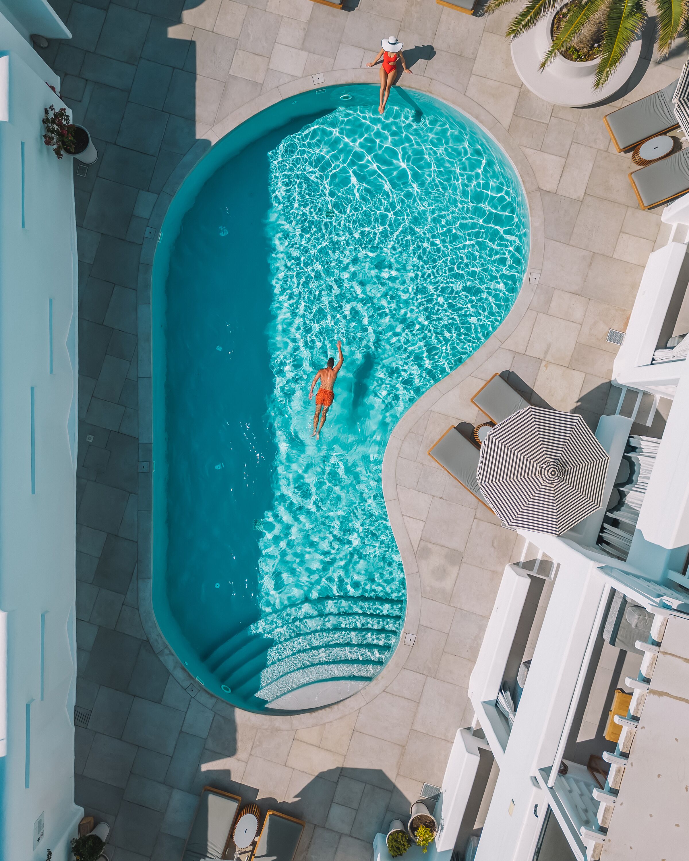 Vista aérea de una piscina con borde curvo, rodeada por un patio empinado con sillas de descanso y sombrillas.