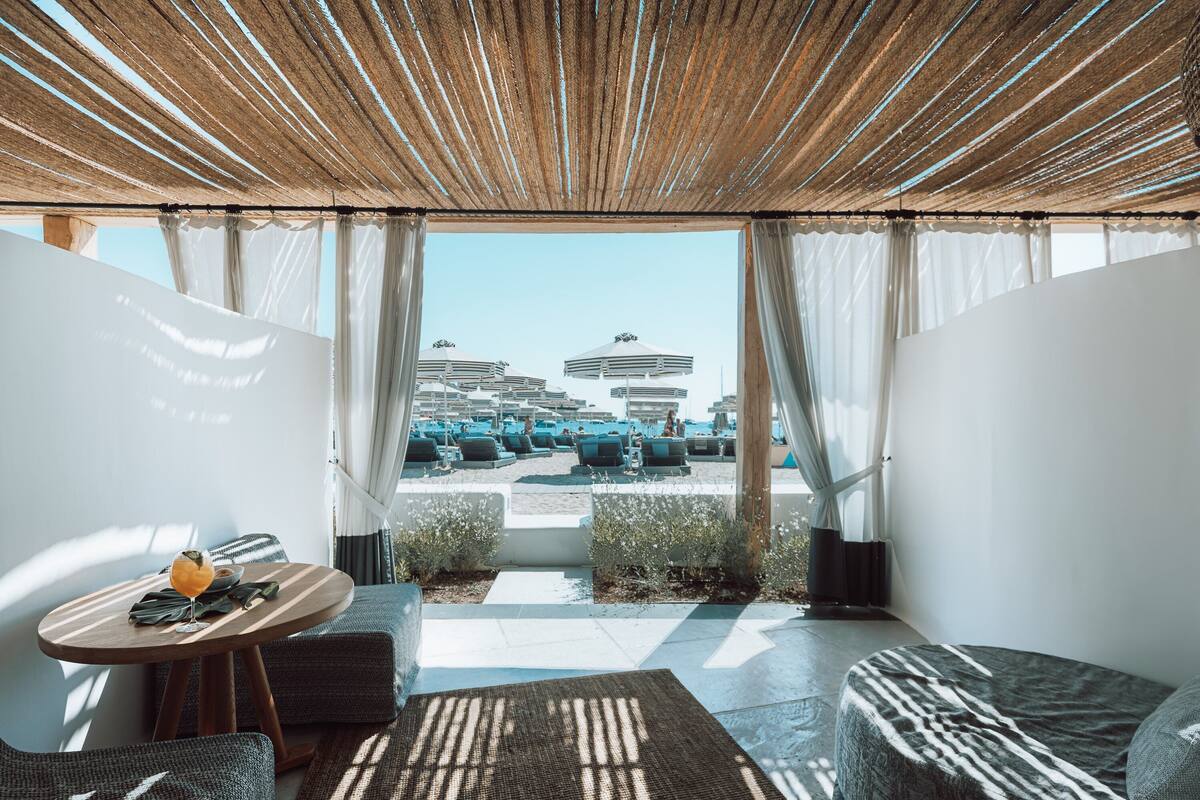 A room with a view of a beach and buildings, featuring a wooden ceiling, a table with a drink, and a sofa.