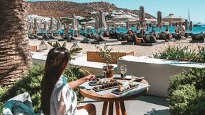 Una mujer cena en una mesa al aire libre con vistas a un resort de playa, que incluye sillas de descanso y sombrillas.