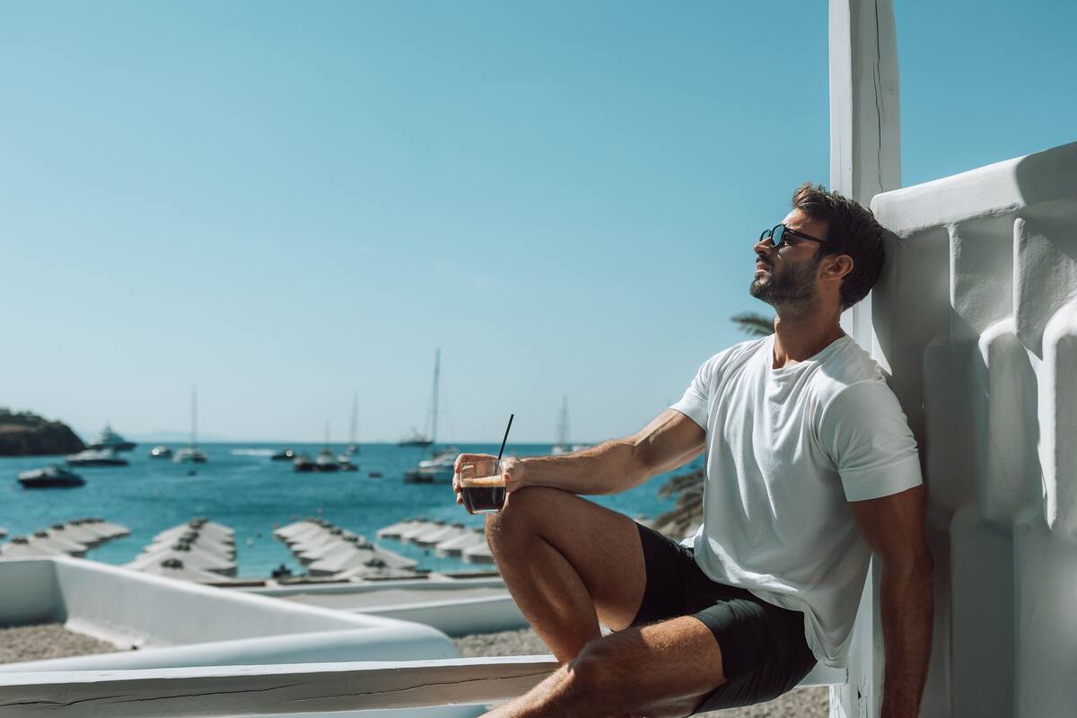 A person sitting by a window overlooking a beach with boats and a clear sky.