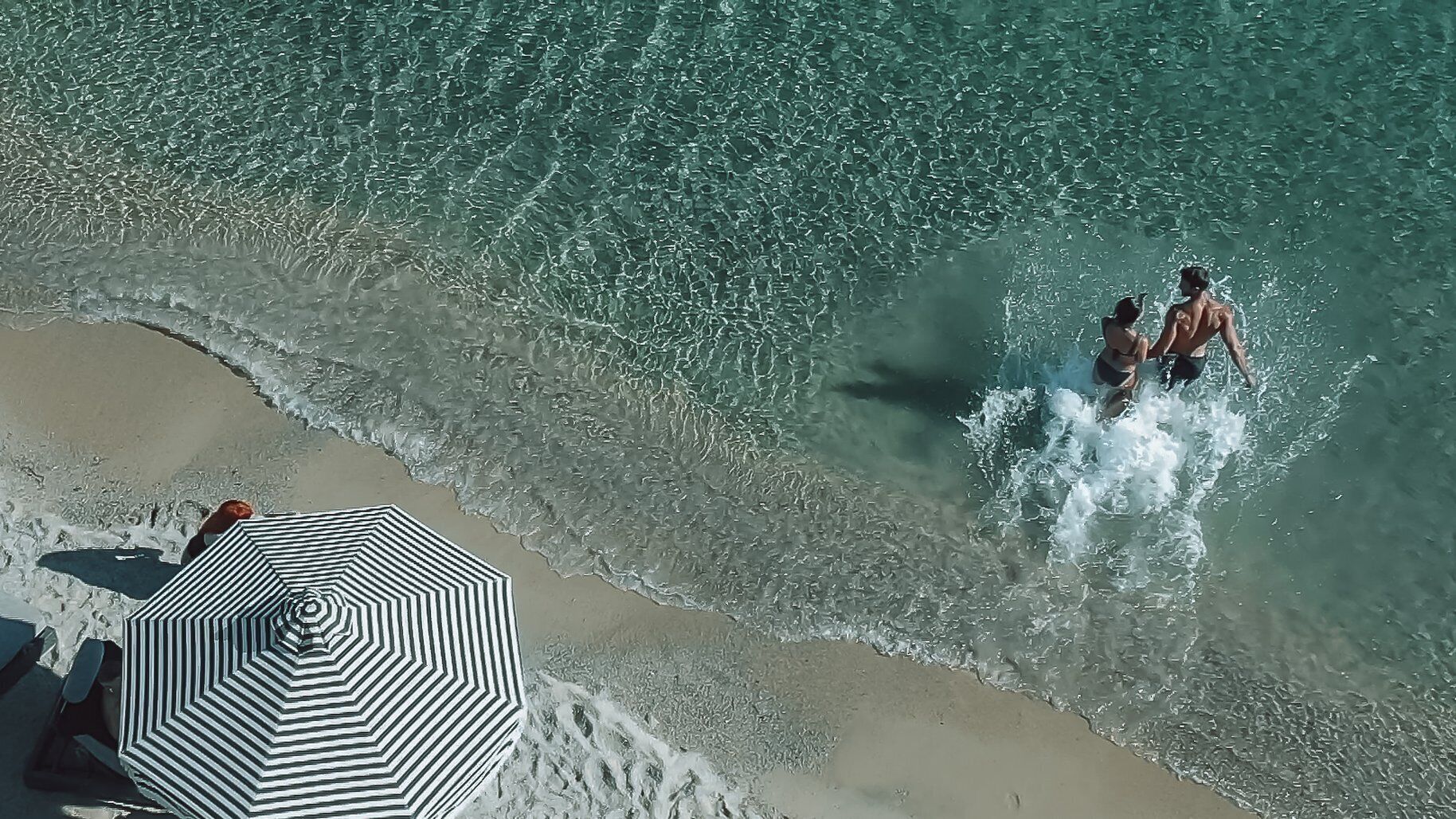 An aerial view of a beach with clear water, a striped umbrella, and people in the water.