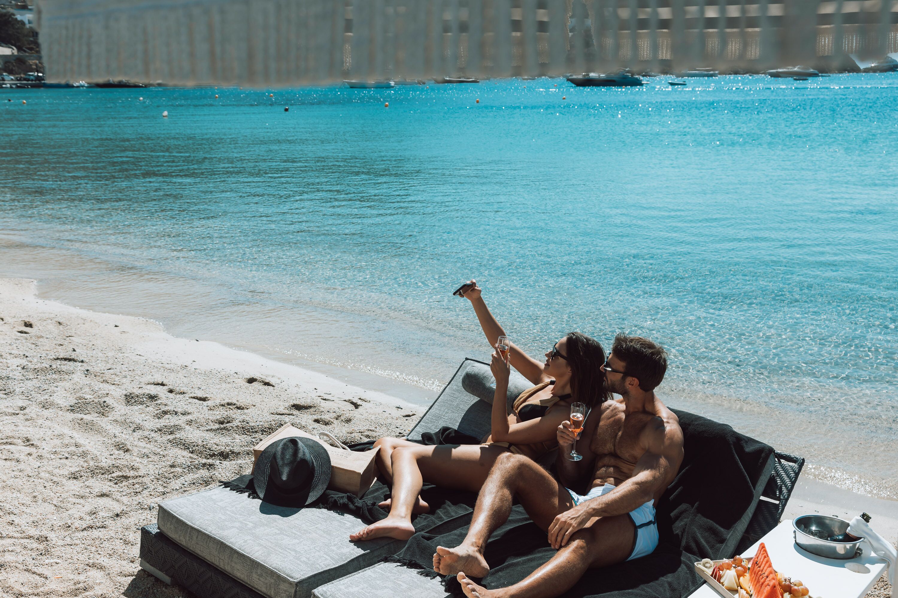 A couple relaxing on a beach chair by the water, with a book and a drink on the table.