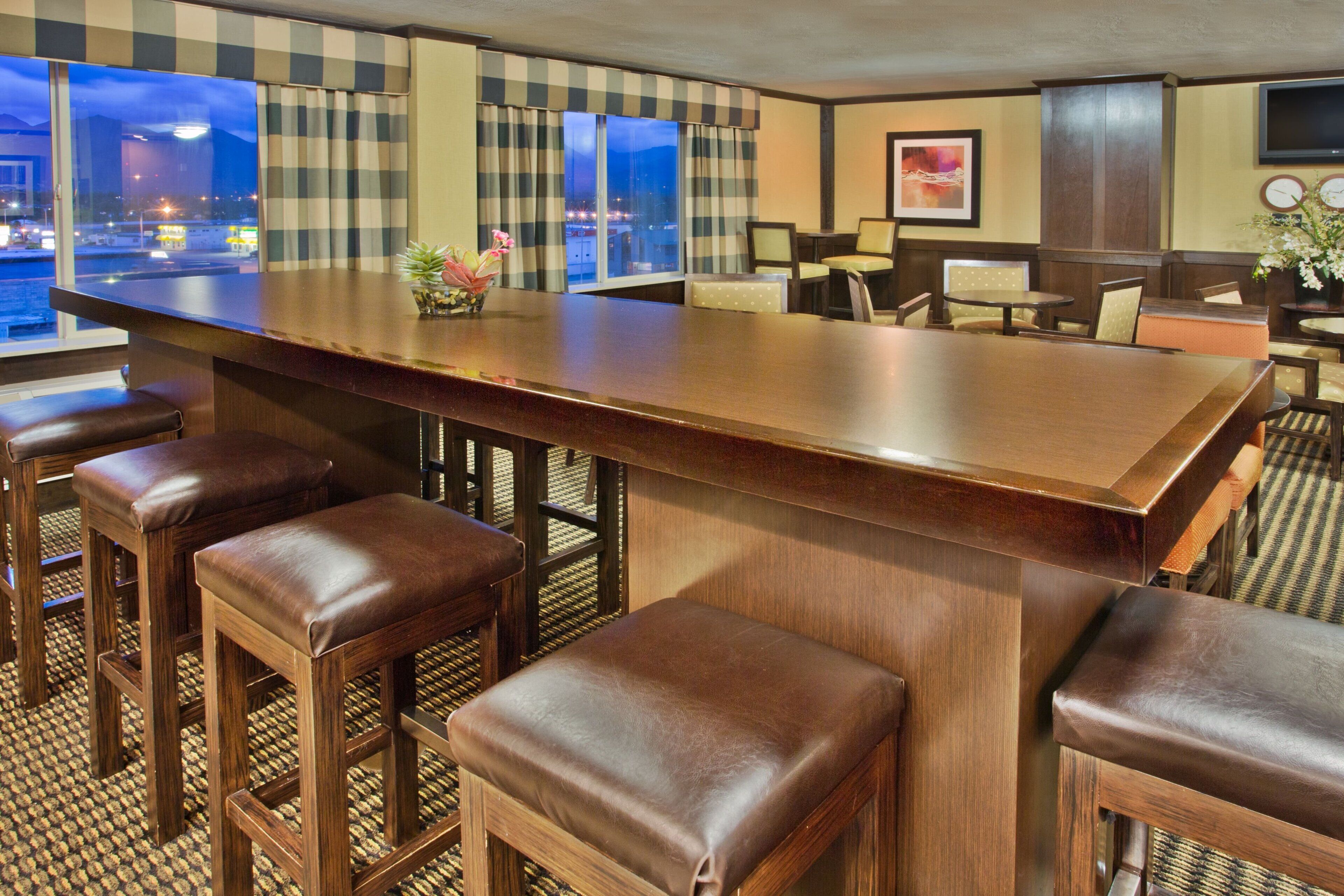 A bar area with a long counter, multiple stools, and a view of the city at night.