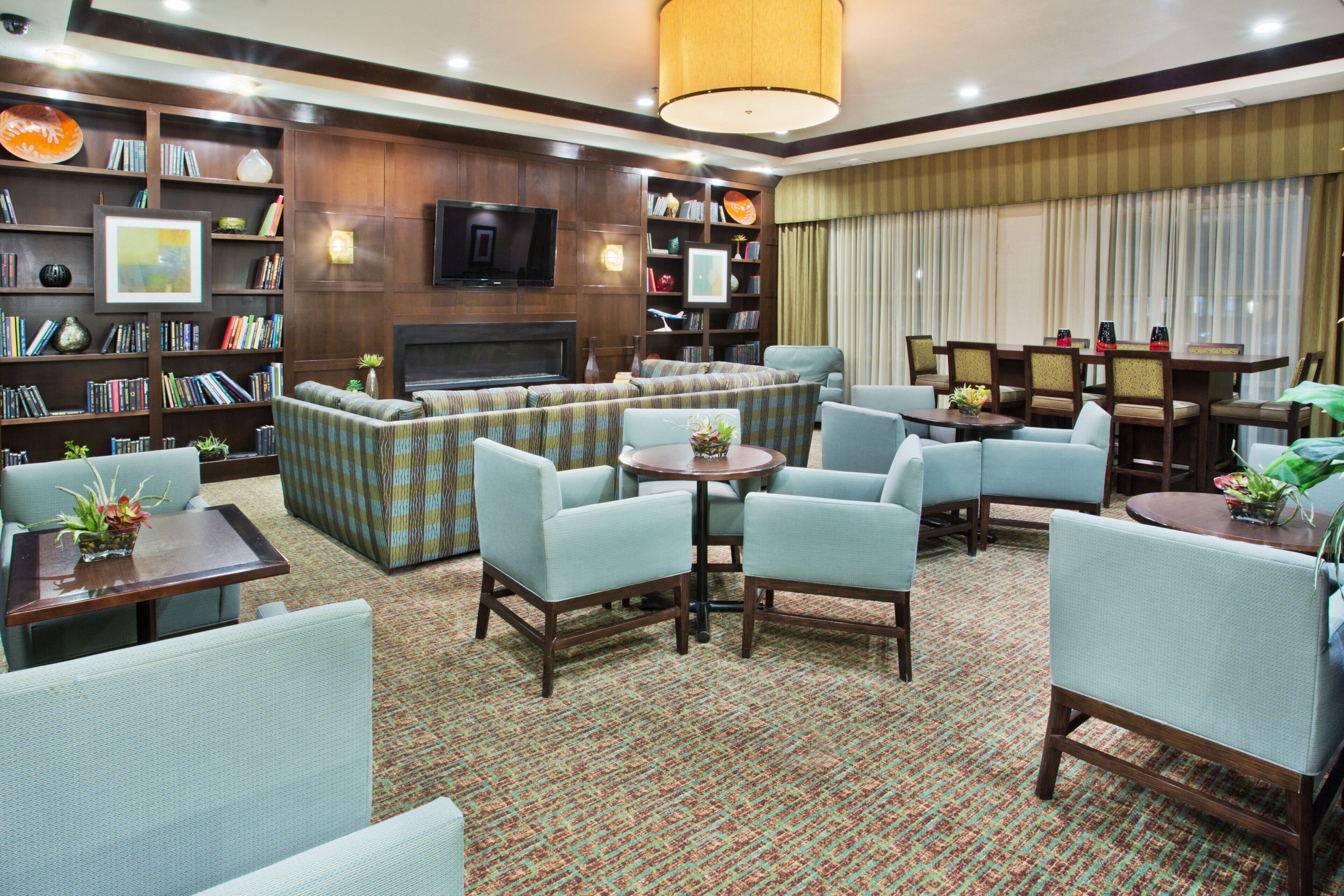 A spacious living area with a sofa set, multiple armchairs, a coffee table, and a fireplace. There are bookshelves filled with books and decorative items, and a television mounted on the wall.
