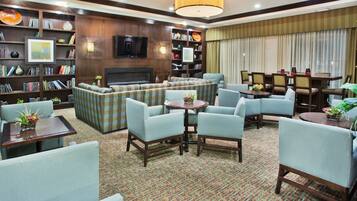 A spacious living area with a sofa set, multiple armchairs, a coffee table, and a fireplace. There are bookshelves filled with books and decorative items, and a television mounted on the wall.