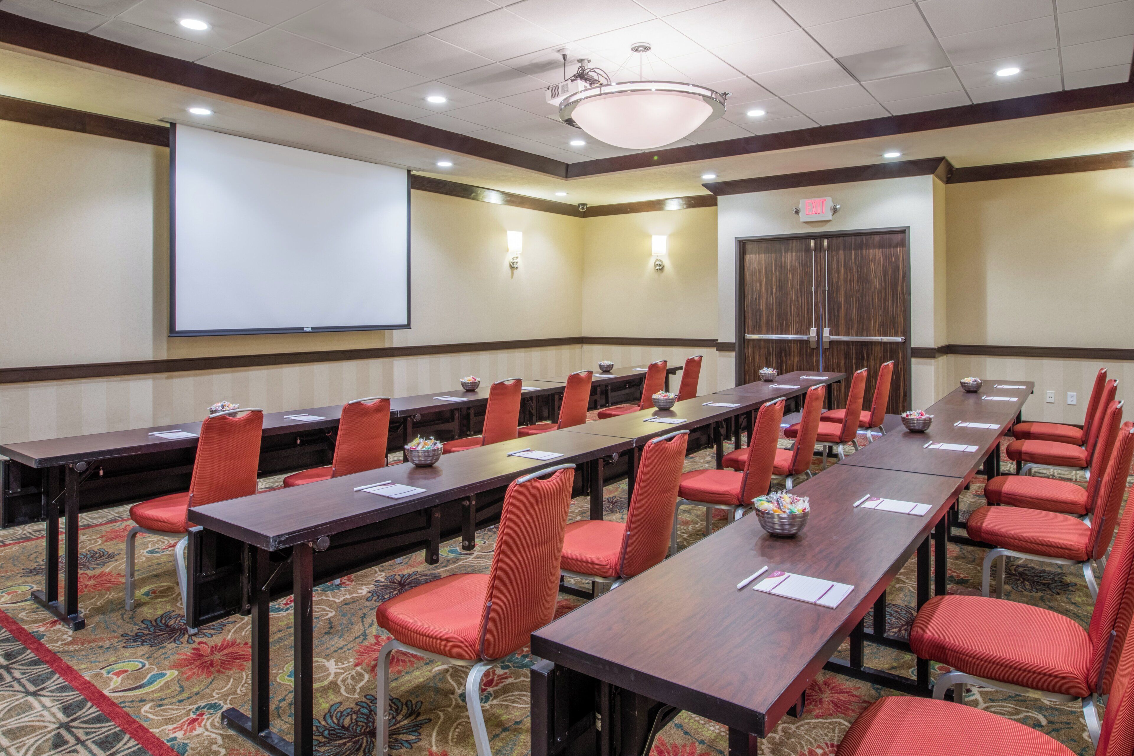A conference room with long tables, red chairs, a projector screen, and an exit sign.
