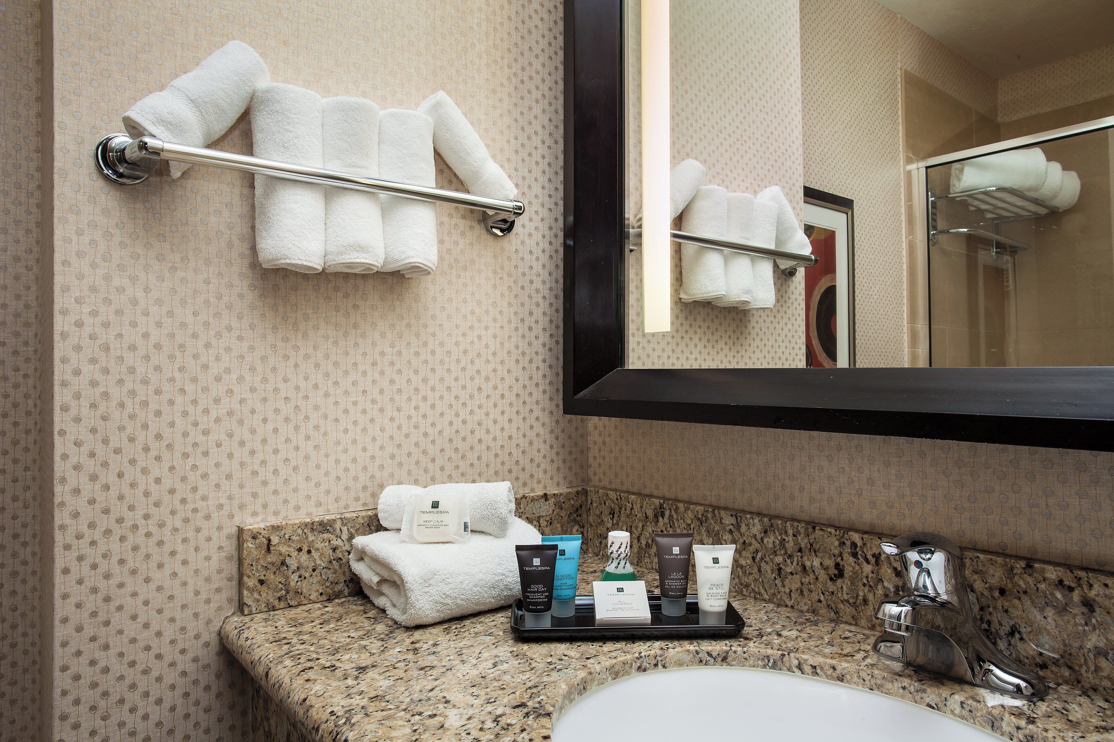 A bathroom with a granite countertop, a sink, and a mirror. There are towels on a towel rack and a tray with toiletries.