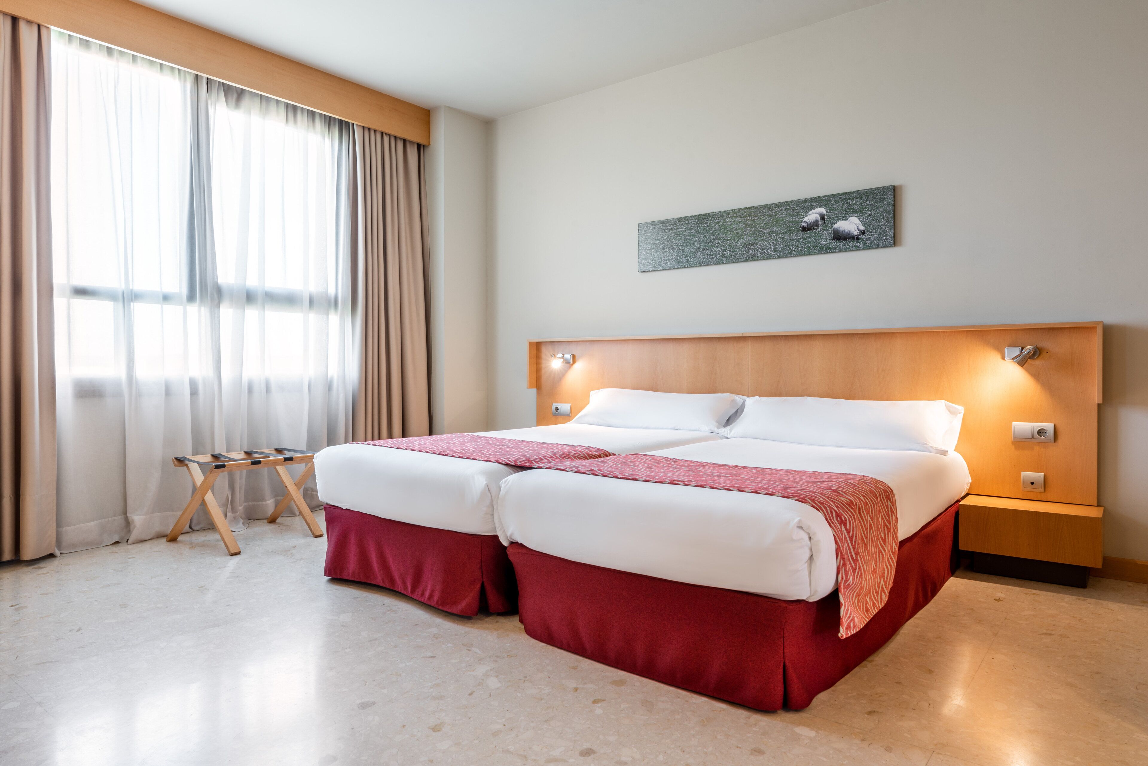 Cama doble con cabecera de madera, mesita de noche pequeña y velador. Se aprecia una ventana con cortinas y un cuadro de ovejas en la pared.