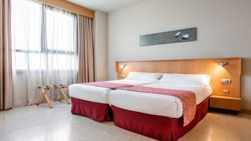 Cama doble con cabecera de madera, mesita de noche pequeña y velador. Se aprecia una ventana con cortinas y un cuadro de ovejas en la pared.