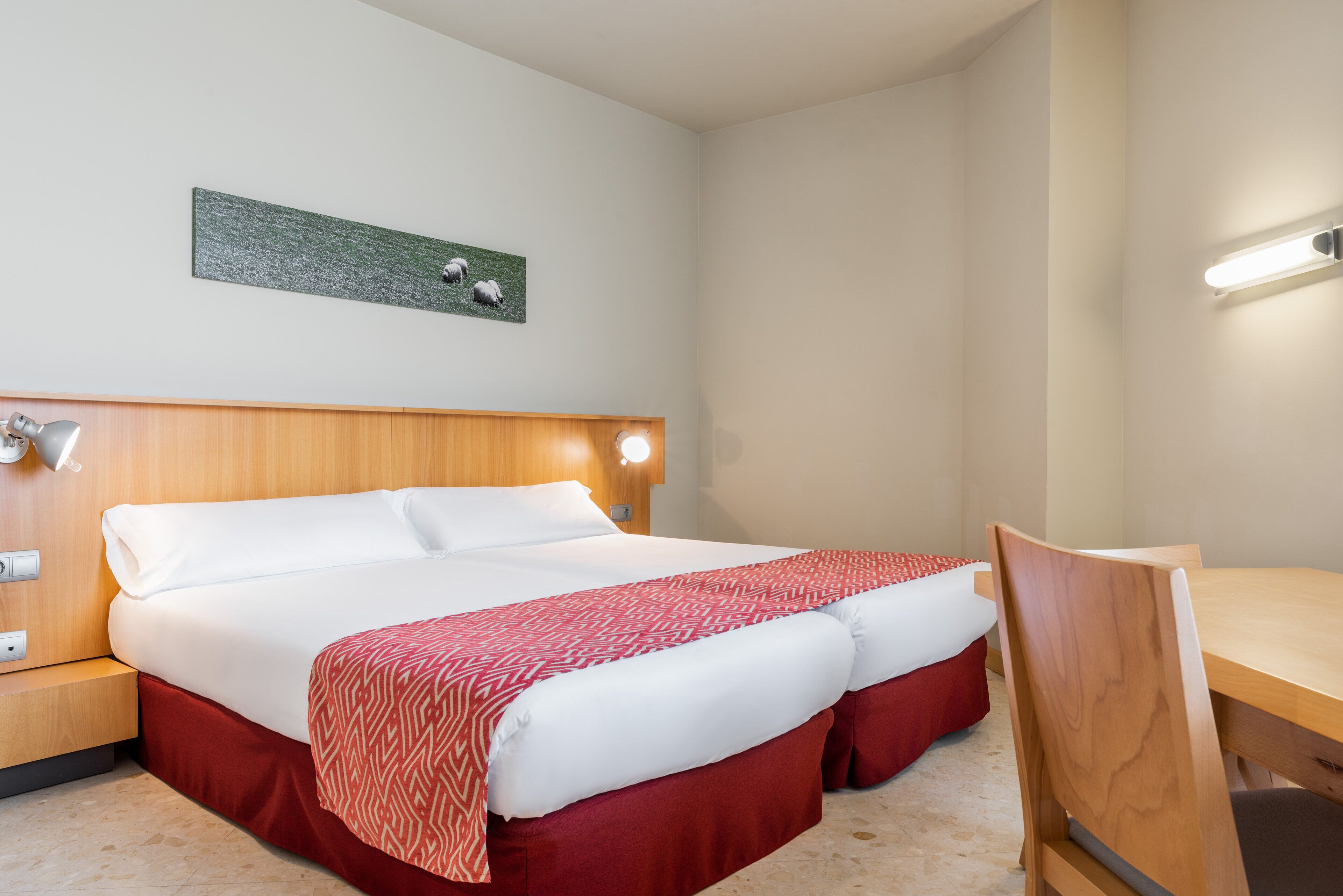 Habitación de hotel con una cama grande, cabecera de madera, lámparas de noche, una mesita y una silla.