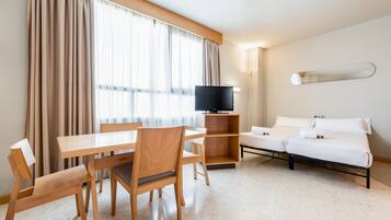 A hotel room with a wooden dining table, chairs, a flat-screen TV, a sofa bed, and a large window with curtains.