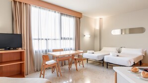 Ein Hotelzimmer mit Fernseher, einem Esstisch mit Stühlen, zwei Betten, einer kleinen Küchenzeile und einem großen Fenster mit Vorhängen.