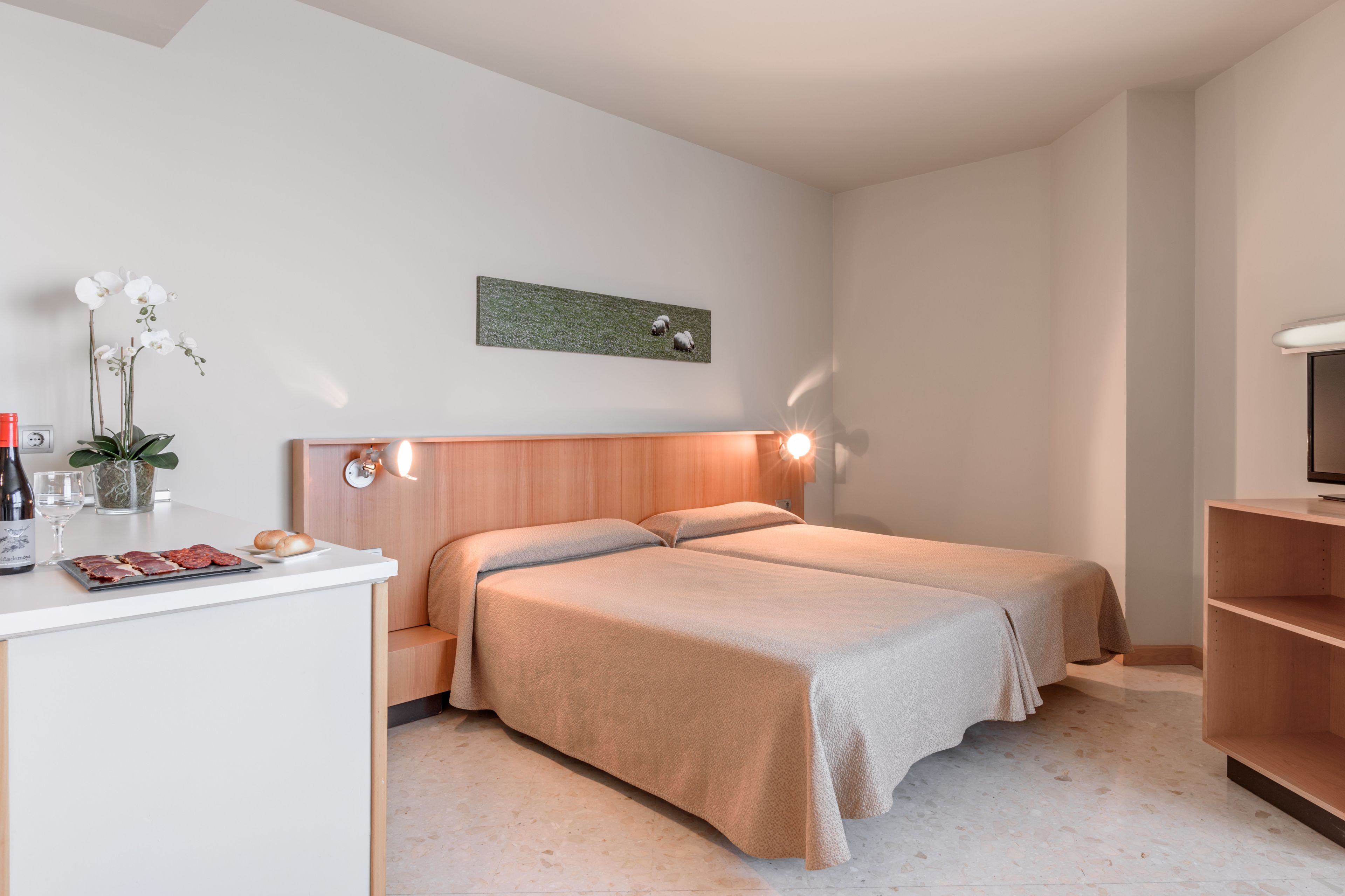 A modern hotel room with two beds, a small desk with a vase of flowers, and a wall-mounted artwork.