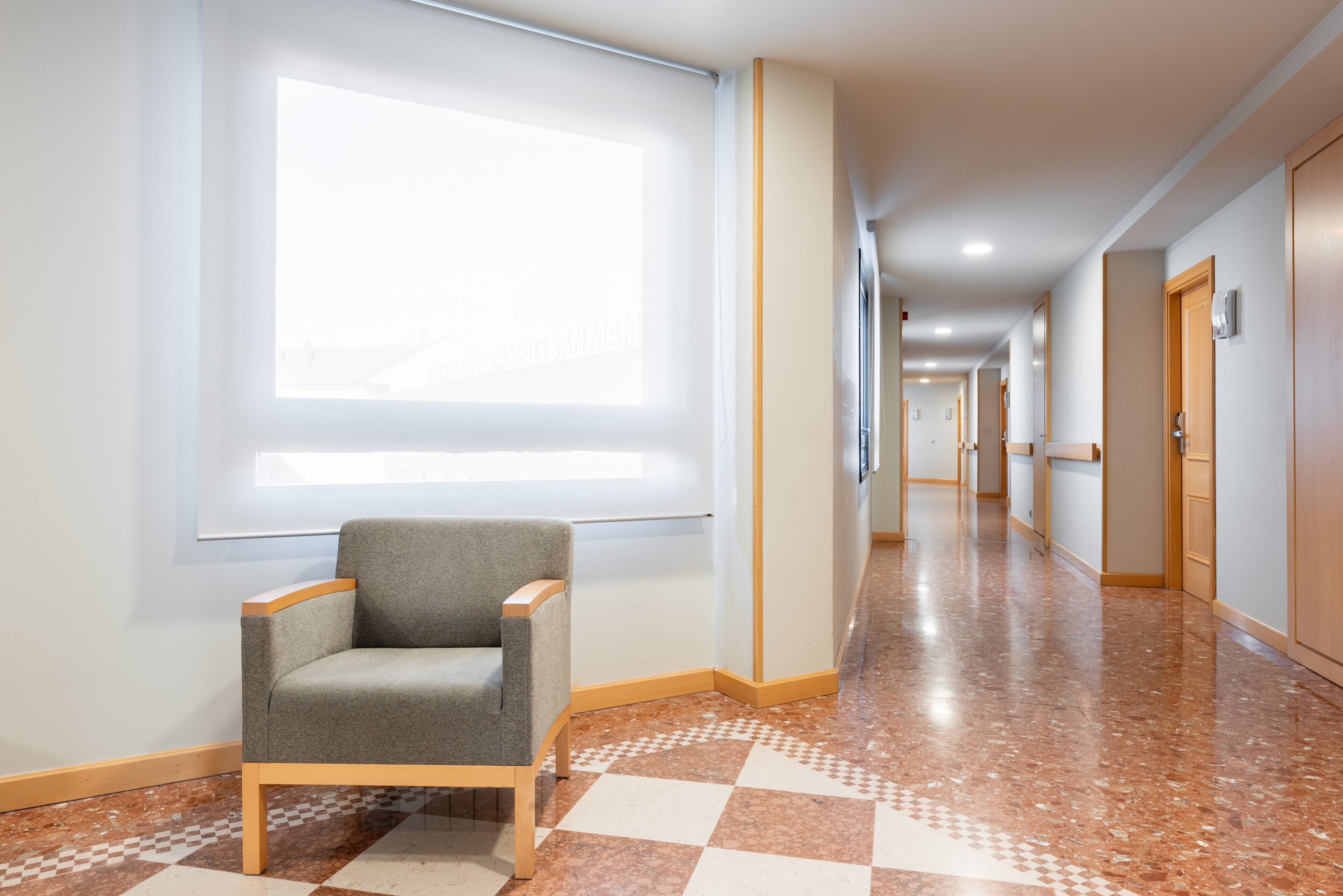 A hotel corridor with a checkered floor, a grey armchair, and a large window providing natural light.