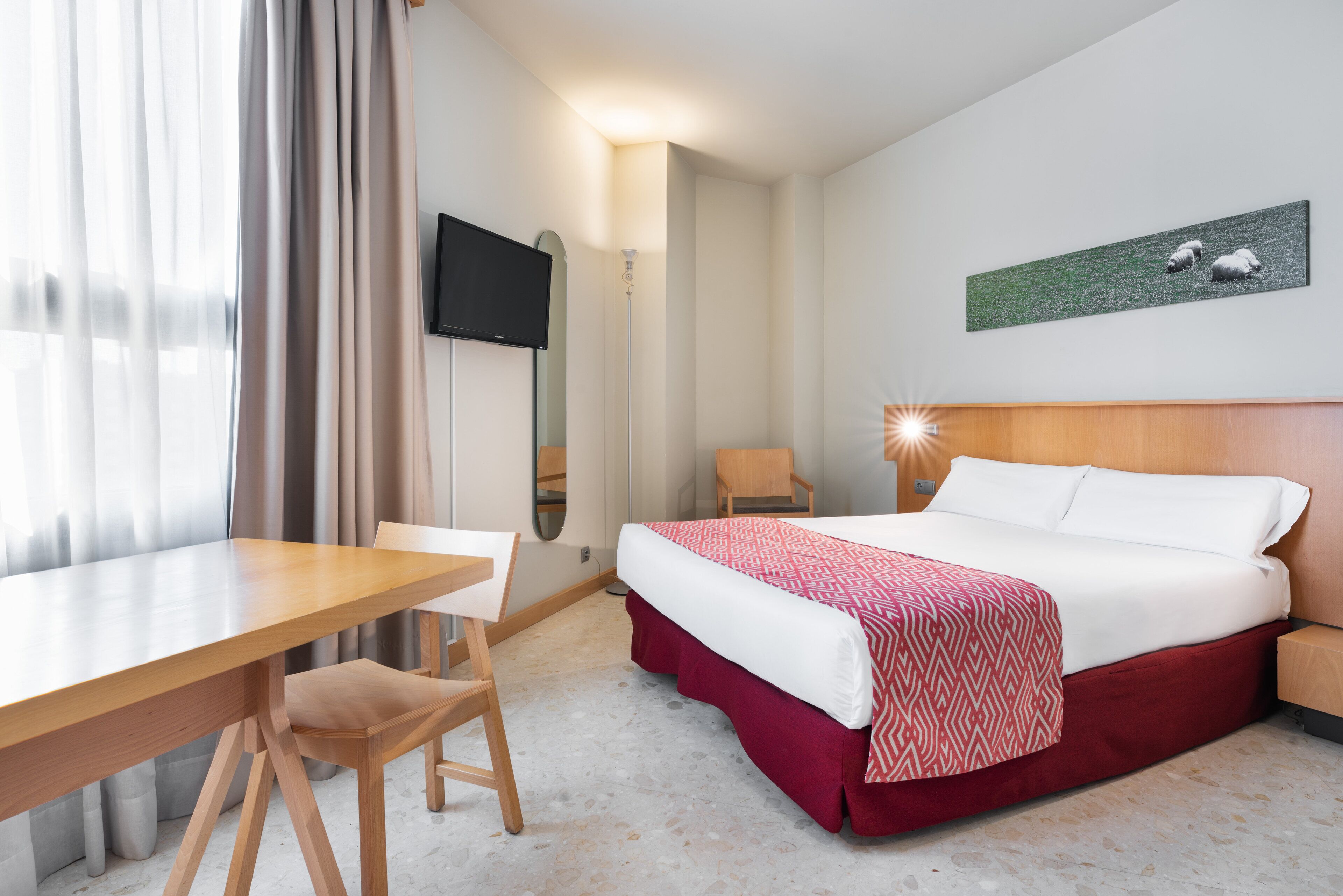 Et hotelværelse med seng, skrivebord, stol, fjernsyn og et billede på væggen.