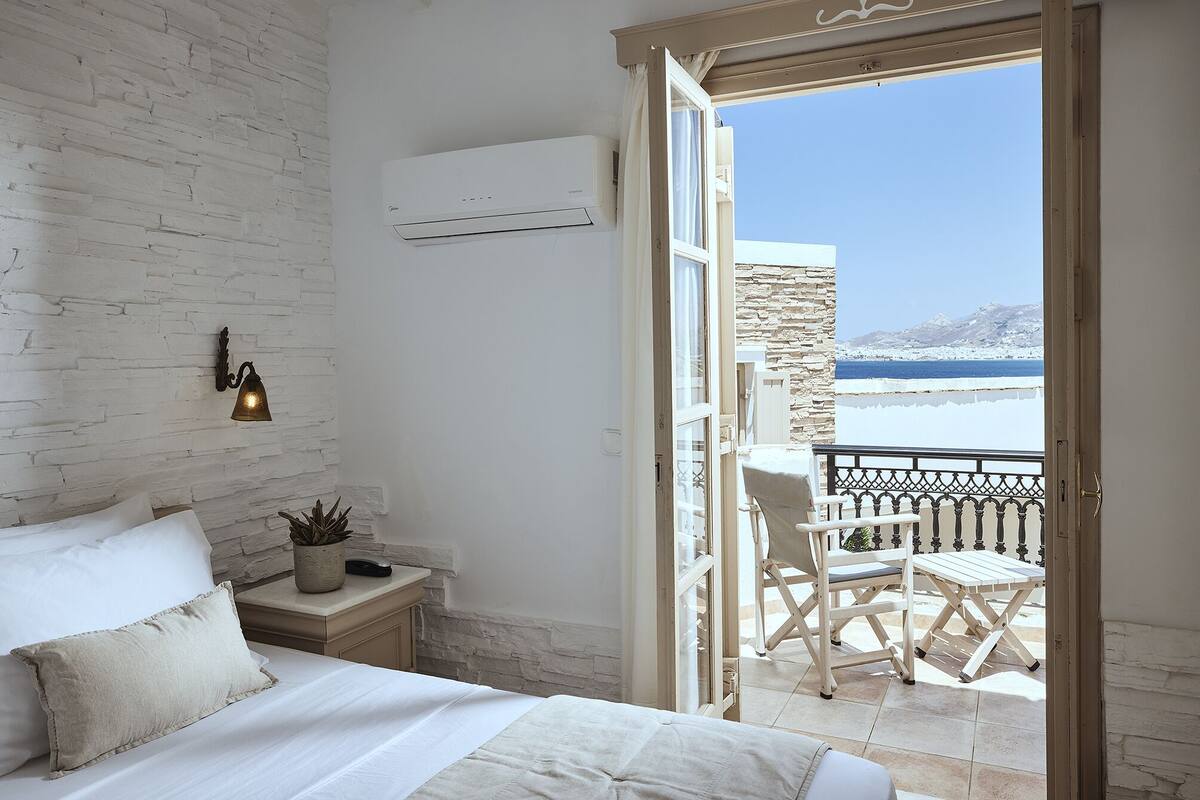 A bedroom with a white brick wall, a bed with white bedding, a bedside table with a lamp, and a view of a balcony with a table and chairs.